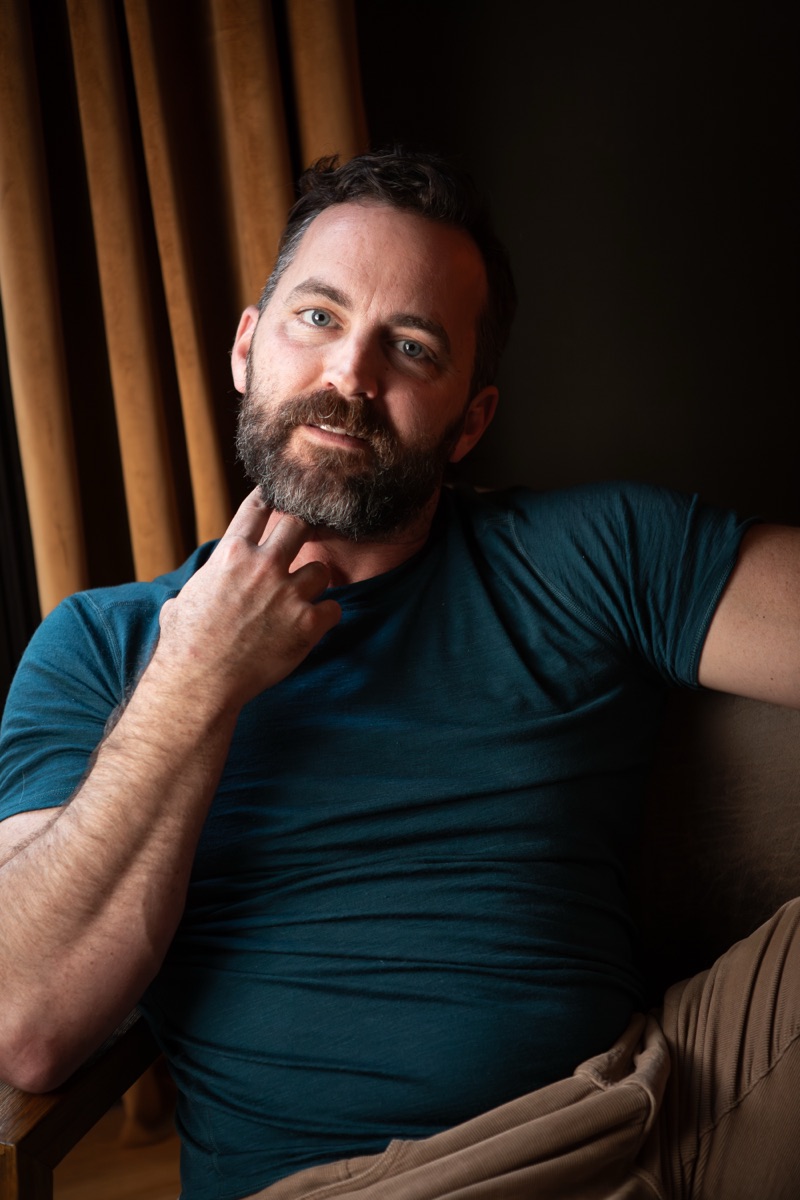 Robert Wilson Smith, actor, seated in a teal T-shirt with a hand at his beard, in front of wooden slats